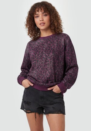 Ung kvinde med krøllet hår iført lilla leopardmønstret sweatshirt og sorte distressed denimshorts, hænderne i lommerne, stående.