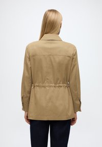 Smėlio spalvos, lengva striukė su siaurinta liemeniu, ilgomis rankovėmis ir užsegamais rankogaliais. Turi klasikinį apykaklę ir lygų paviršių.