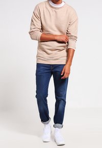 Krémová mikina s kulatým výstřihem a dlouhými rukávy, spárovaná s tmavě modrými džínami a bílými sportovními teniskami. Hladká textura látky a volný střih.