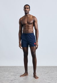 Námořnické modré plavkové šortky s elastickým pasem a stahovací šňůrkou, bočními kapsami a červenou nášivkou s logem na levé noze.