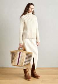 Kremowy sweter z dzianiny z prążkowanym wykończeniem, beżowa spódnica midi z rozcięciem z przodu, brązowe botki oraz pleciona torba na ramię w paski.