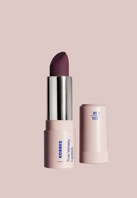 Korres True Velvety Lipstick in una tonalità di prugna profonda, con una texture opaca, contenuta in un astuccio cilindrico rosa chiaro con dettagli argentati.