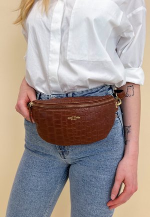 Marsupio in pelle marrone con motivo a coccodrillo, dotato di hardware dorato, chiusura con zip superiore e una tracolla regolabile, indossato in vita.