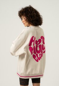 Krém színű pulóver, laza szabással, amelynek hátsó részén merész rózsaszín "HOT MUM CLUB" grafika található, és a hemnél rózsaszín csíkos részletekkel van díszítve.