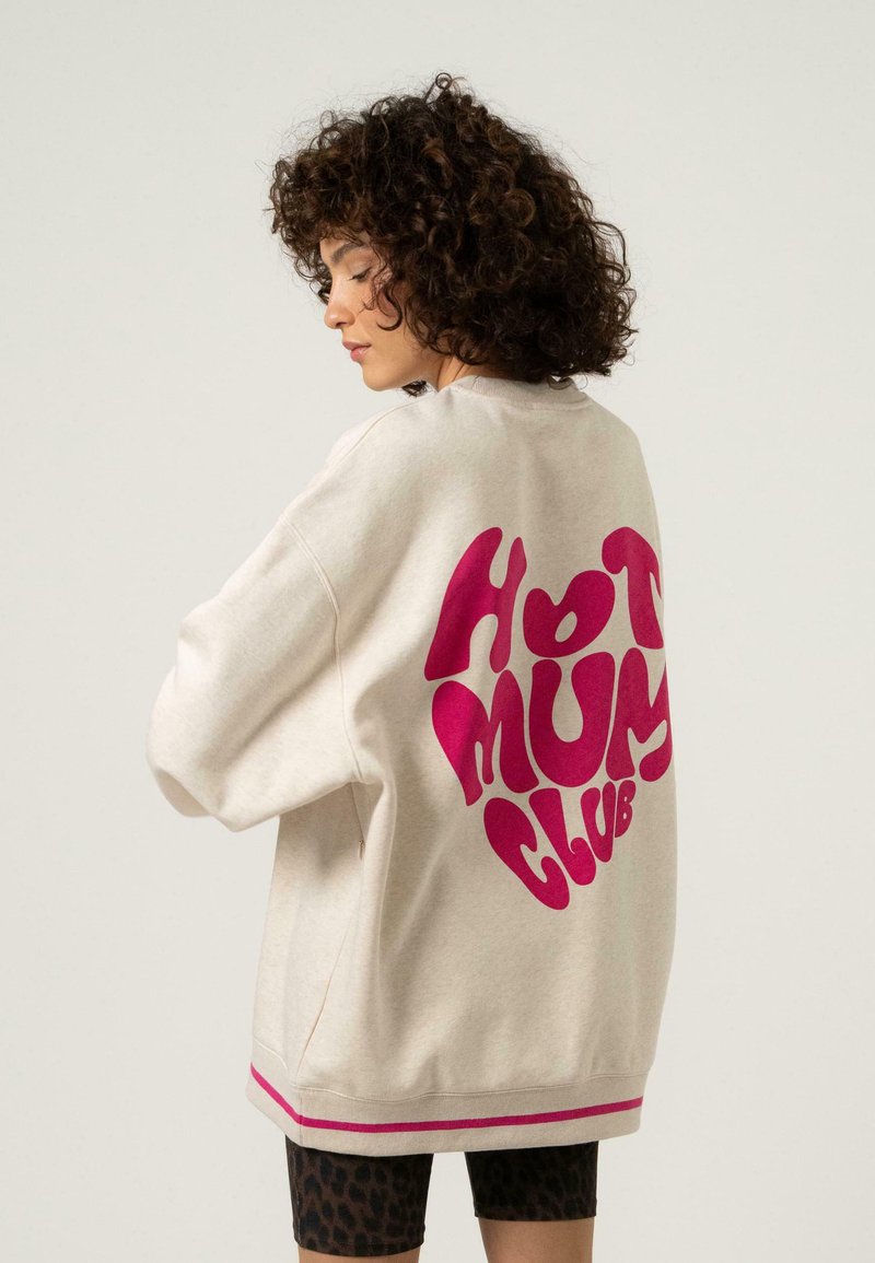 Krém színű pulóver, laza szabással, amelynek hátsó részén merész rózsaszín "HOT MUM CLUB" grafika található, és a hemnél rózsaszín csíkos részletekkel van díszítve.