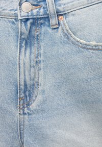 Los jeans de mezclilla azul claro presentan costuras visibles, un botón de metal y un ligero desgaste cerca de la zona de los bolsillos, con un diseño de cinco bolsillos.