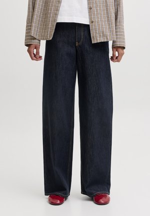 Personne portant un jean large foncé, des chaussures plates rouges, une chemise blanche et une veste à carreaux marron, debout devant un fond uni.