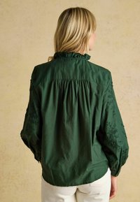 Dunkelgrüne Baumwollbluse mit Rüschenausschnitt, gerafft im Rücken und bestickten Blumenmotiven an den Ärmeln. Sie hat lange, puffige Ärmel.