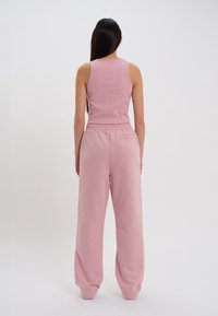 Donna con lunghi capelli scuri che indossa una maglietta senza maniche rosa a costine e pantaloni rosa larghi, in piedi su uno sfondo bianco.