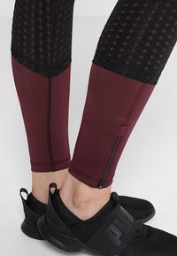 Zbliżenie na dolne nogi w bordowych legginsach z czarną siateczką i detalami w postaci zamka, w połączeniu z czarnymi butami na slip-on marki Puma.