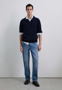 Suéter de punto en navy con un cuello ancho, sobre una camisa de rayas azul claro; combinado con jeans azul claro y zapatos negros.
