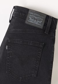 Črne denim hlače z pravokotno, blagovno označeno nalepko na pasu. Imajo zadnji žep z značilnimi šivi.
