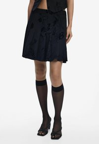 Jupe plissée noire avec un motif floral, associée à des chaussettes noires transparentes montantes avec des bandes solides, et des sandales noires à talons ouverts.