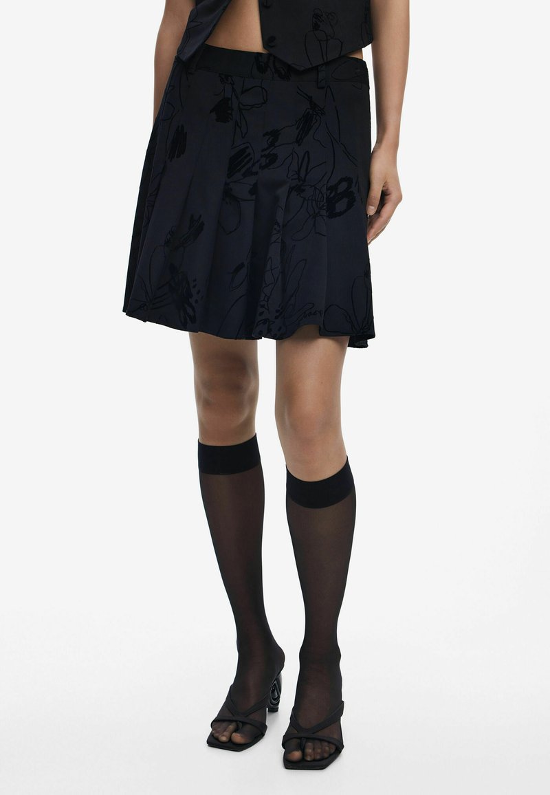 Jupe plissée noire avec un motif floral, associée à des chaussettes noires transparentes montantes avec des bandes solides, et des sandales noires à talons ouverts.