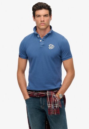 Blauwe polo met korte mouwen, geborduurd logo op de borst, gecombineerd met donkere spijkerbroeken en een rood geruit shirt om de taille geknoopt.