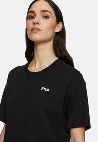 Black cotton t-shirt with short sleeves, round neckline, and a white Fila logo on the chest. Smooth texture, casual fit.