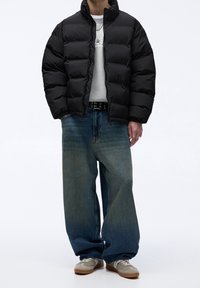 Personne portant une doudoune noire, un sweat-shirt gris clair, un jean large bleu, des baskets grises et des chaînes argentées, debout devant un fond blanc.