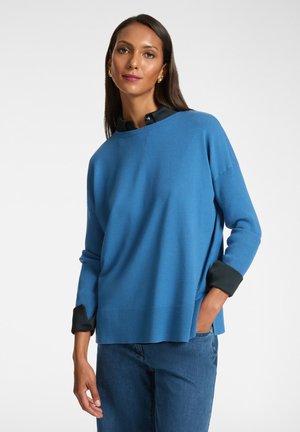 Suéter de punto azul con un corte relajado, acentos acanalados en las mangas y el dobladillo, con una camisa negra con cuello debajo. Textura simple y suave.