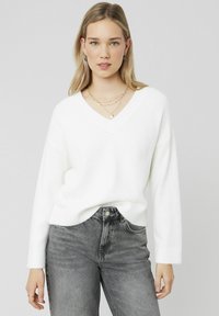 Pull en V blanc en matière douce avec des manches longues, associé à un jean taille haute gris. Design simple, détails minimaux, coupe décontractée.