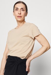 T-shirt bege de manga curta com decote redondo, textura suave e corte descontraído. Combinado com uma saia preta com nó na cintura.