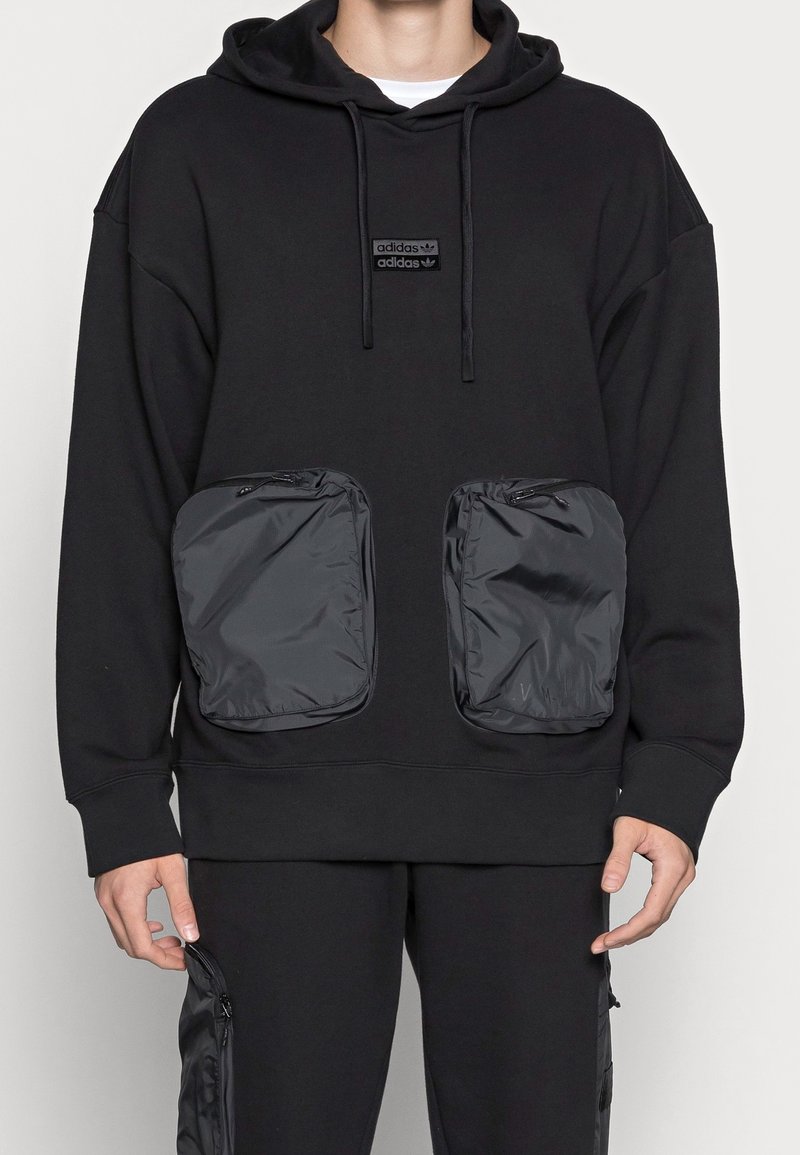Person som bär en svart Adidas-hoodie med stora dragkedjefickor i nylon fram och matchande svarta byxor med sidofickor med dragkedja.