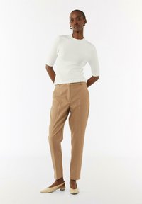 Kobieta stoi z rękami za plecami, ubrana w białą bluzkę z krótkim rękawem, beżowe dopasowane spodnie i kremowe buty na niskim obcasie.