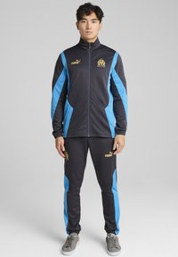 Puma träningsoverall i mörkgrått och blått. Innehåller en zip-up jacka med sidostripes och logotyp, matchande byxor med elastiska muddar.