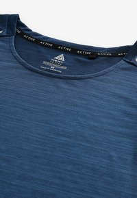 Granatowa koszulka sportowa z teksturowanej tkaniny, okrągłym dekoltem i wzmocnionymi szwami na ramionach. Metka głosi "NEXT ACTIVE" oraz "WYPRODUKOWANO W KAMBODŻY."