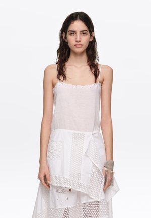 Giovane donna con lunghi capelli scuri che indossa un vestito bianco senza maniche con applicazioni in pizzo e un braccialetto argentato con frange al polso destro.