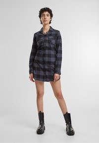Vestito camicia a scacchi nero e blu con maniche lunghe, chiusura con bottoni frontale, due tasche sul petto e una vestibilità leggermente sagomata. Abbinato a stivali neri alla caviglia.