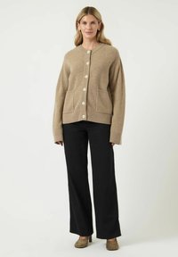 Beige vlněný cardigan s kulatým výstřihem, velkými knoflíky a dvěma předními kapsami, v kombinaci s černými kalhotami s širokými nohavicemi a taupe podpatky.