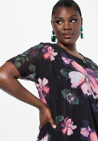 Blusa nera floreale con fiori rosa e viola, maniche corte e decorazioni. Modella che indossa orecchini a statement verdi.