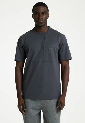 Man draagt een donkergrijs shirt met korte mouwen met een klein logo op de borst en lichtgrijze broek, staand tegen een effen witte achtergrond.