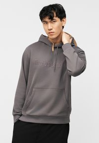 Νέος άνδρας που φοράει γκρι φούτερ Timberland με τσέπη μπροστά, αγγίζοντας τον λαιμό του, σε απλό φωτεινό φόντο.