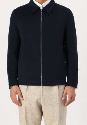 Hombre con chaqueta azul marino con cremallera y cuello, sobre una camisa blanca y pantalones plisados beige, de pie frente a un fondo liso.