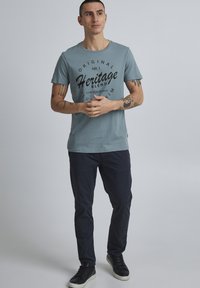 Svijetloplava pamučna majica s crnim grafičkim printom, okrugli izrez, kombinirana s tamnoplavim hlačama i crnim tenisicama. Model stoji s rukama sklopljenim.