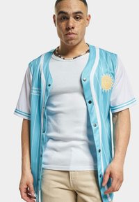 Tricou sportiv de culoare albastră deschisă cu dungi verticale albe, având un logo cu soare. Purtați peste un maieu alb cu nervuri, asortat cu pantaloni bej.