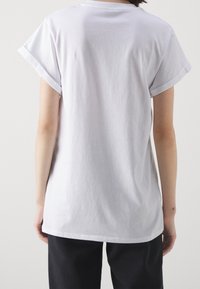 Bílý bavlněný tričko s ohrnutými krátkými rukávy, volný střih, jednolitá textura a bez viditelných vzorů nebo grafik. Pohled zezadu.