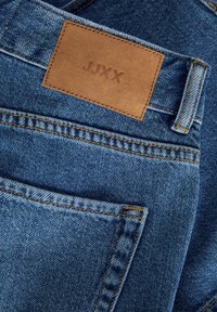 Poche arrière d'un jean en denim bleu avec une étiquette de marque en cuir marron cousue au-dessus, affichant le texte "JJXX".