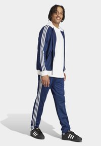 Marineblå træningsdragter med hvide striber ned ad ærmerne og benene. Har en lynlåsjakke foran og tætsiddende bukser, parret med sorte sneakers.