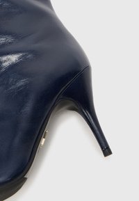 Námornícky modrá kožená topánka na vysokom opätku s špicatou špičkou a stiletto opätkom. Obsahuje lesklú textúru a minimálnu kovovú výzdobu.
