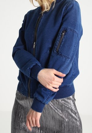 Person iført en blå lynlåstrøje med en lynlåslomme på armen og et metallisk plisseret nederdel, stående med armene krydset.