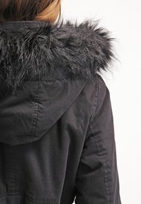 Osoba w czarnej kurtce z kapturem wykończonym czarnym sztucznym futrem, pokazana z tyłu na tle białym.