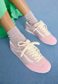 Baskets roses et blanches avec un design en daim et maille, dotées de lacets épais couleur crème et de chaussettes roses texturées sur une surface bleue.