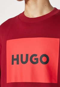 Homme portant un sweat-shirt rouge à col rond avec un grand rectangle orange et le texte noir gras "HUGO" sur la poitrine.
