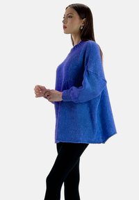 Maglione lavorato a maglia oversize viola con maniche lunghe e a sbuffo e una texture a coste. L'orlo è leggermente più lungo sul retro, abbinato a leggings neri.