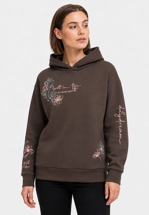 Vrouw draagt een bruine hoodie met bloemenpatronen en tekst "lost in the moment," "daydream," en "full wonder" op de mouwen en borst.