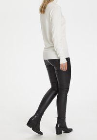 Weißer Rollkragenpullover kombiniert mit schwarzen, veganen Lederleggings und Stiefeletten. Der Pullover hat gerippte Bündchen und eine taillierte Silhouette.