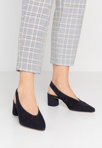 Námořnicky modré semišové boty na podpatku se zavazováním za patu, špičaté špičky a masivní podpatky, s výrazným hlubokým V-říznutím.