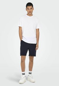 Camiseta blanca de algodón combinada con pantalones cortos de textura azul marino. Usada con zapatillas blancas y calcetines a rayas en blanco y negro. Ajuste casual.